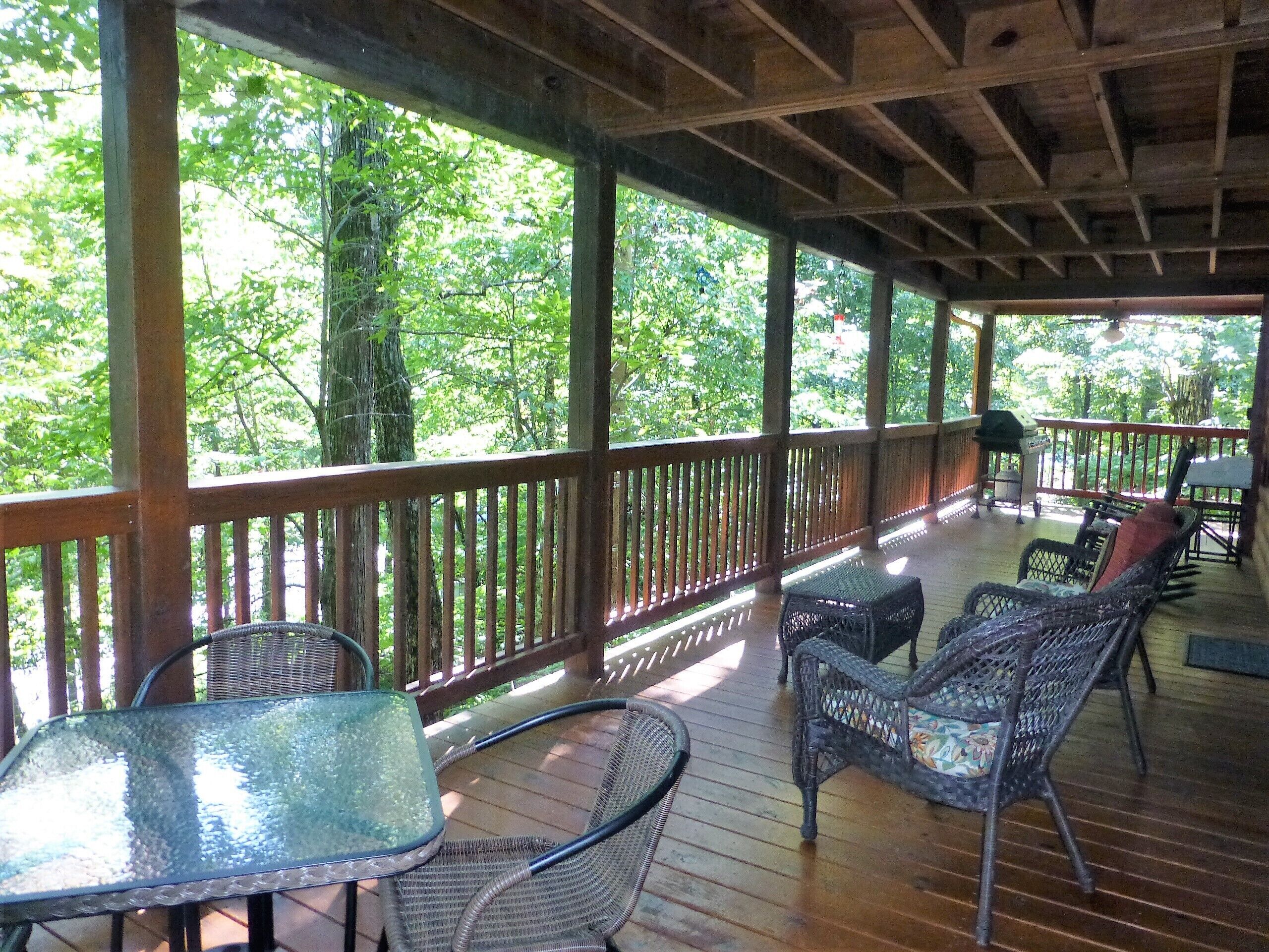 A covered outdoor deck with wooden railings, wicker furniture, and a glass table.