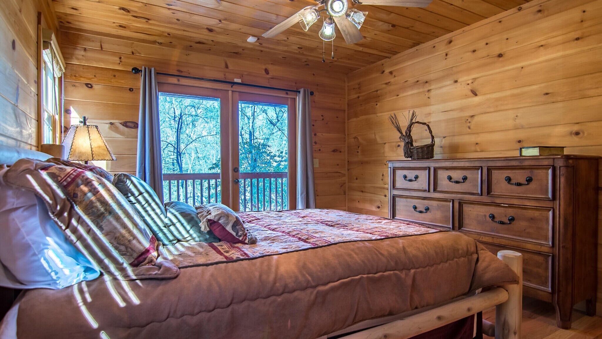 A wooden cabin bedroom with a bed, a dresser, a ceiling fan, and a window with a view of trees.
