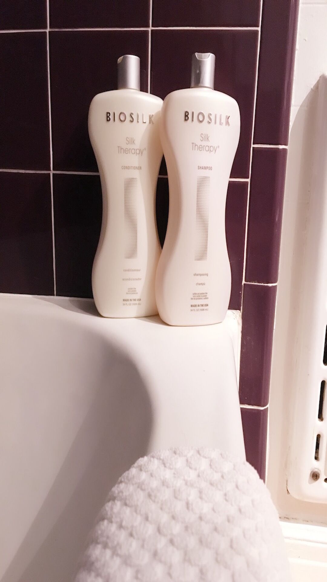 Due flaconi di balsamo e shampoo Biosilk Silk Therapy su una superficie bianca, con un muro rivestito di piastrelle sullo sfondo.