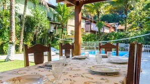 An outdoor dining area with a table set for two, surrounded by lush greenery and a pool in the background.