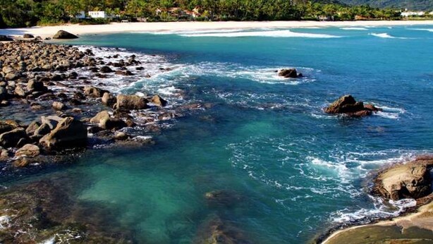 A coastal scene with clear turquoise water, rocky shoreline, and a distant beach with buildings.