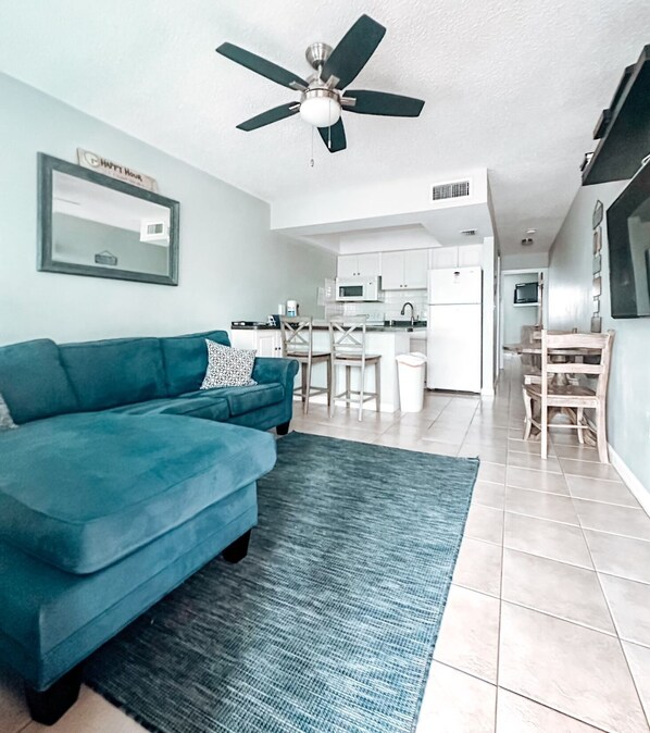 A modern living room with a blue sofa, a ceiling fan, and a kitchen in the background.