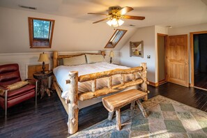 Une chambre à coucher avec un lit en bois, un fauteuil rouge, un banc en bois, un ventilateur de plafond et une fenêtre donnant sur des arbres.