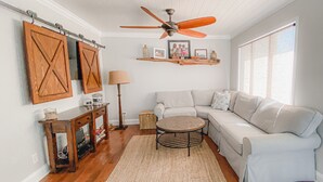 Ein Wohnzimmer mit Ecksofa, rundem Couchtisch, Deckenventilator, Holztüren im Scheunenstil und einem Fenster mit transparenten Vorhängen.
