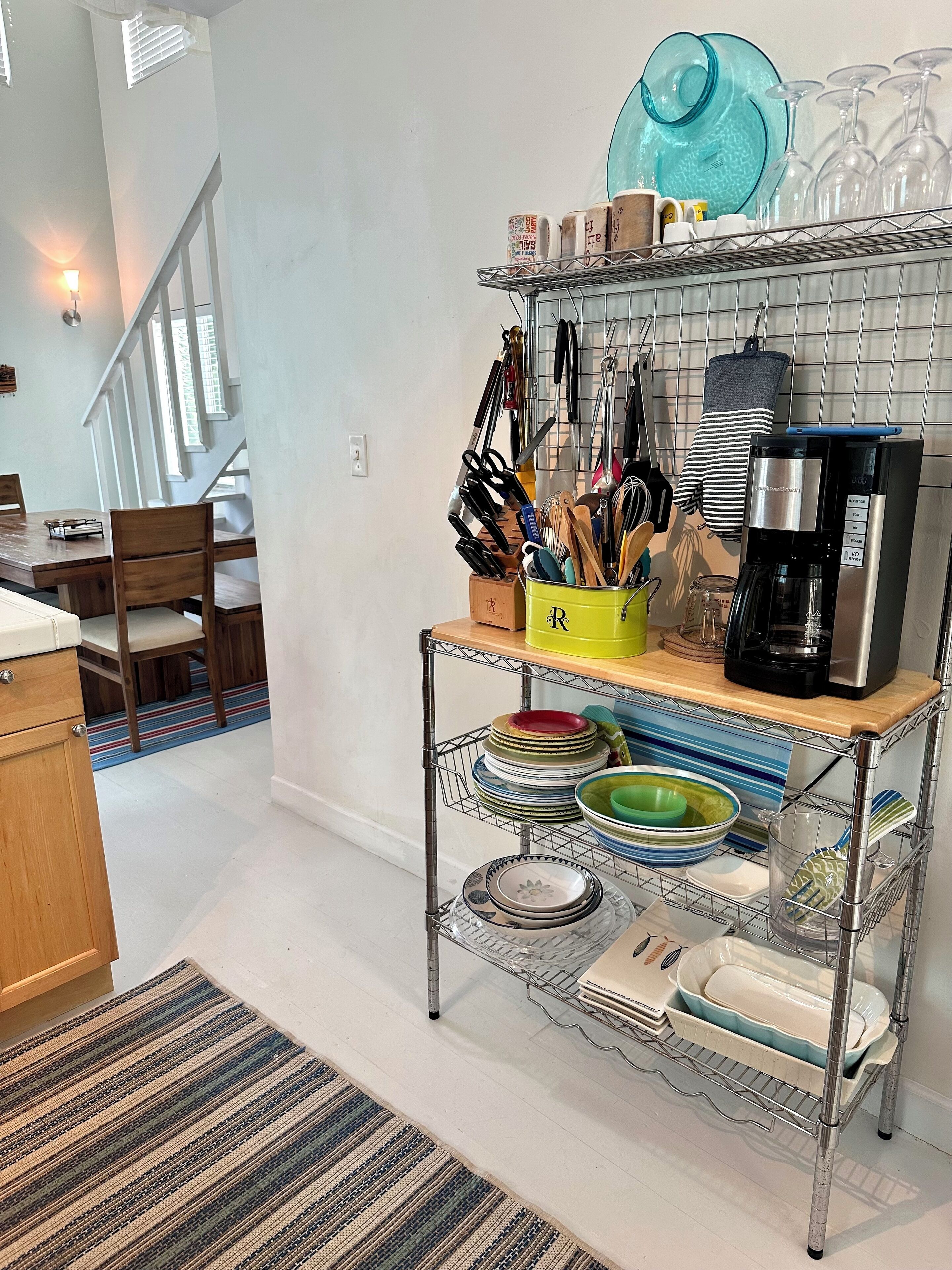 A kitchen with a metal rack holding plates, bowls, and a coffee maker. There is a dining table with chairs and a striped rug.