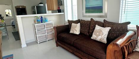 Ein Wohnzimmer mit einem braunen Sofa, einem Holzschaukelstuhl und einem Streifen Teppich. Im Hintergrund ist eine Küchenzeile mit einem Kühlschrank und einer Mikrowelle zu sehen.