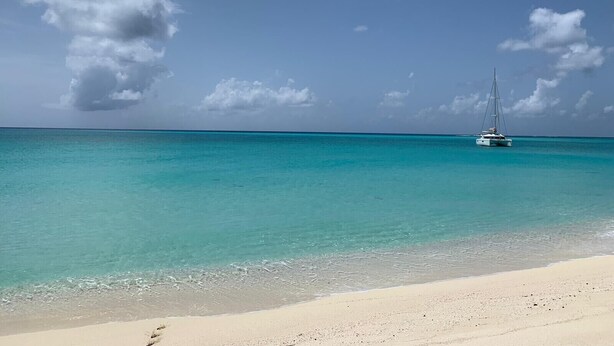 Een strand met helder turquoise water en een zeilboot in de verte.