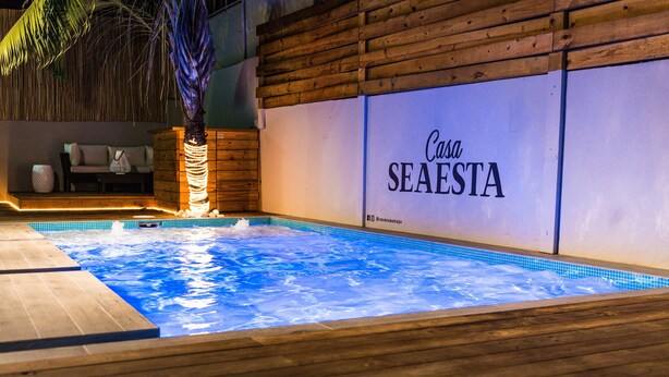 Una piscina con cubierta de madera, un área de descanso con sofá y cojines, y un letrero que dice "Casa SEAESTA".