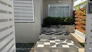 Una entrada moderna al aire libre con un camino de baldosas a cuadros que lleva a una puerta, rodeada por una valla de madera y follaje.