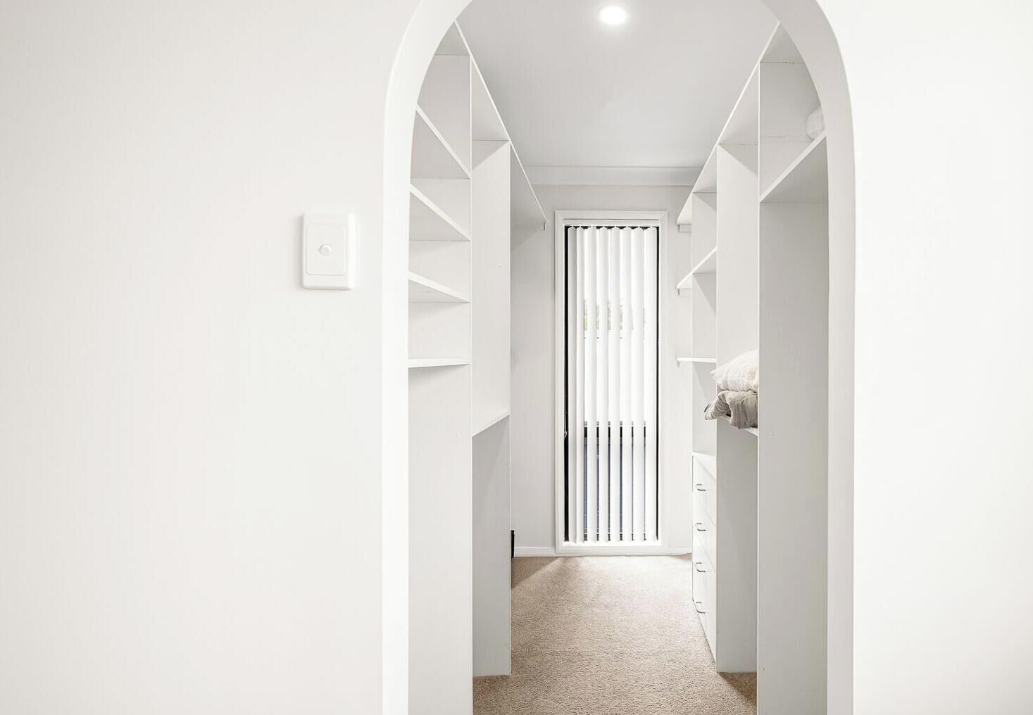 Een smalle gang met witte muren en een tapijten vloer, die leidt naar een kamer met een bed en een raam met verticale blinds.
