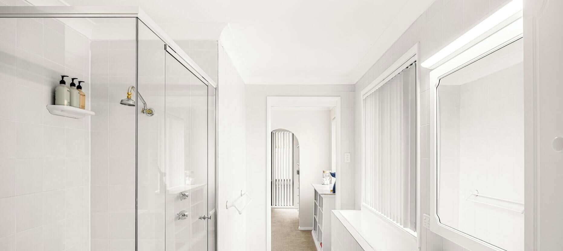 Un baño moderno con cabina de ducha de vidrio, pared con azulejos blancos y estante empotrado con artículos de aseo.