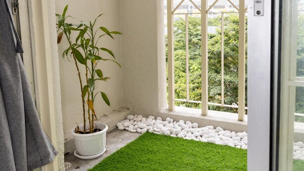 A balcony with a small patch of artificial grass, a potted plant, and a view of a cityscape through a barred window.