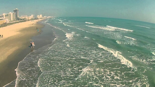 A beach with a sandy shore and ocean waves. There are buildings in the background.