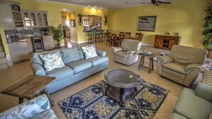 A spacious living room with a grey sofa, beige armchairs, and a coffee table. A dining area with wooden chairs and a kitchen with cabinets and a sink are visible in the background.
