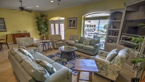 A spacious living room with a large sofa, two armchairs, a coffee table, and a bookshelf. There is a large window with a view of the ocean and a balcony.