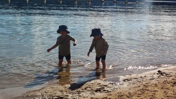 Two children playing in shallow water near a sandy beach with a clear sky and distant mountains.