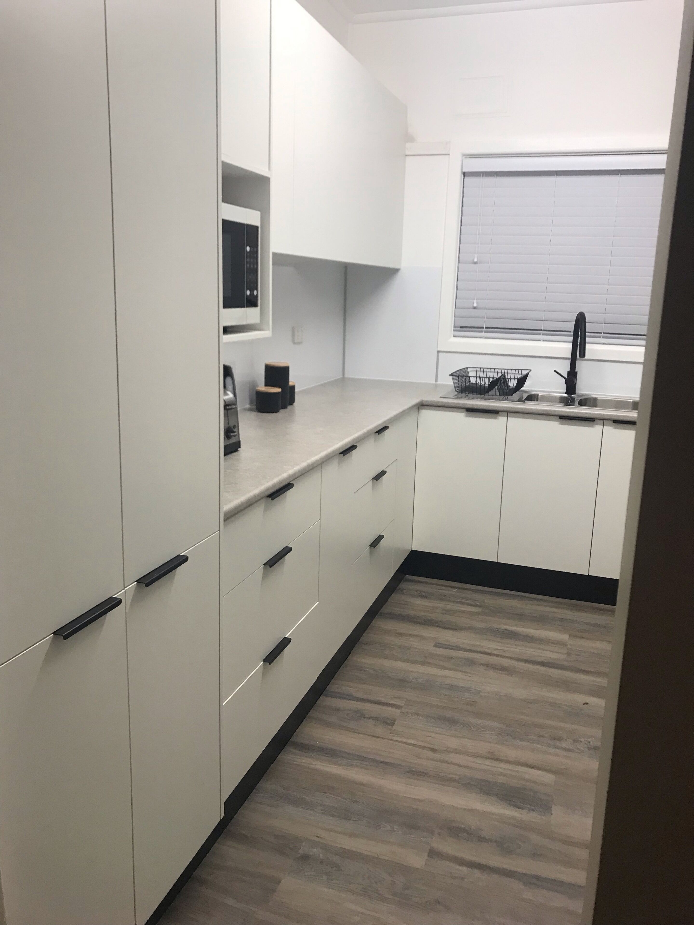 A modern kitchen with white cabinets, a black countertop, and a window with blinds.