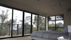 Quarto com cama grande, ventilador de teto e vista para árvores por grandes janelas.