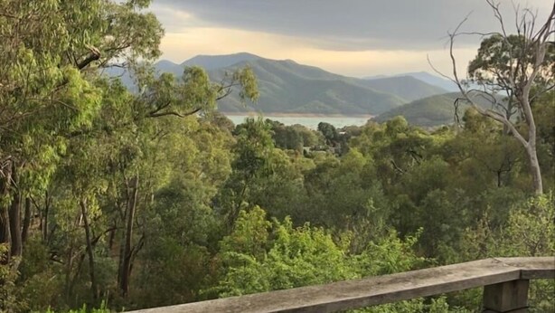 Vista de um deck de madeira com guarda-corpo de metal, com vista para uma floresta densa e montanhas ao longe.