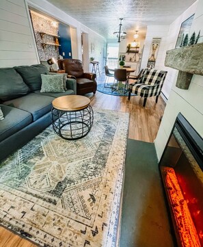 Un salon chaleureux avec un canapé, un fauteuil, une table basse et une cheminée. On aperçoit à l’arrière-plan un coin repas avec des chaises et une table.