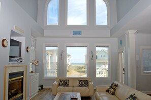 A spacious living room with a fireplace, a television, and a large window overlooking a beach.