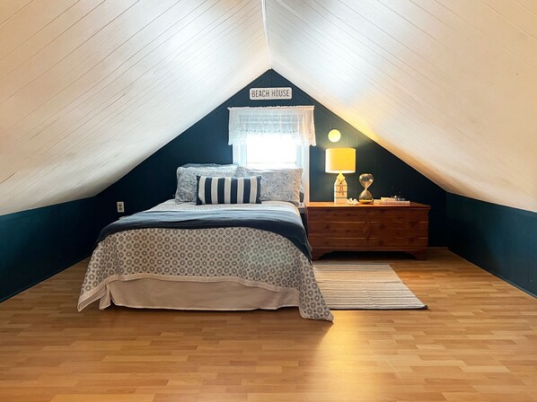 A bedroom with a sloped ceiling, a bed with patterned bedding, a nightstand with a lamp, and a sign reading "BEACH HOUSE".