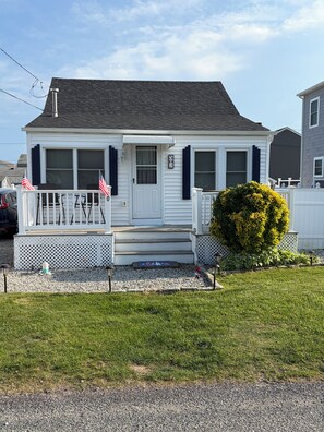 A single-story house with a white exterior, a front porch with a railing, and two American flags.