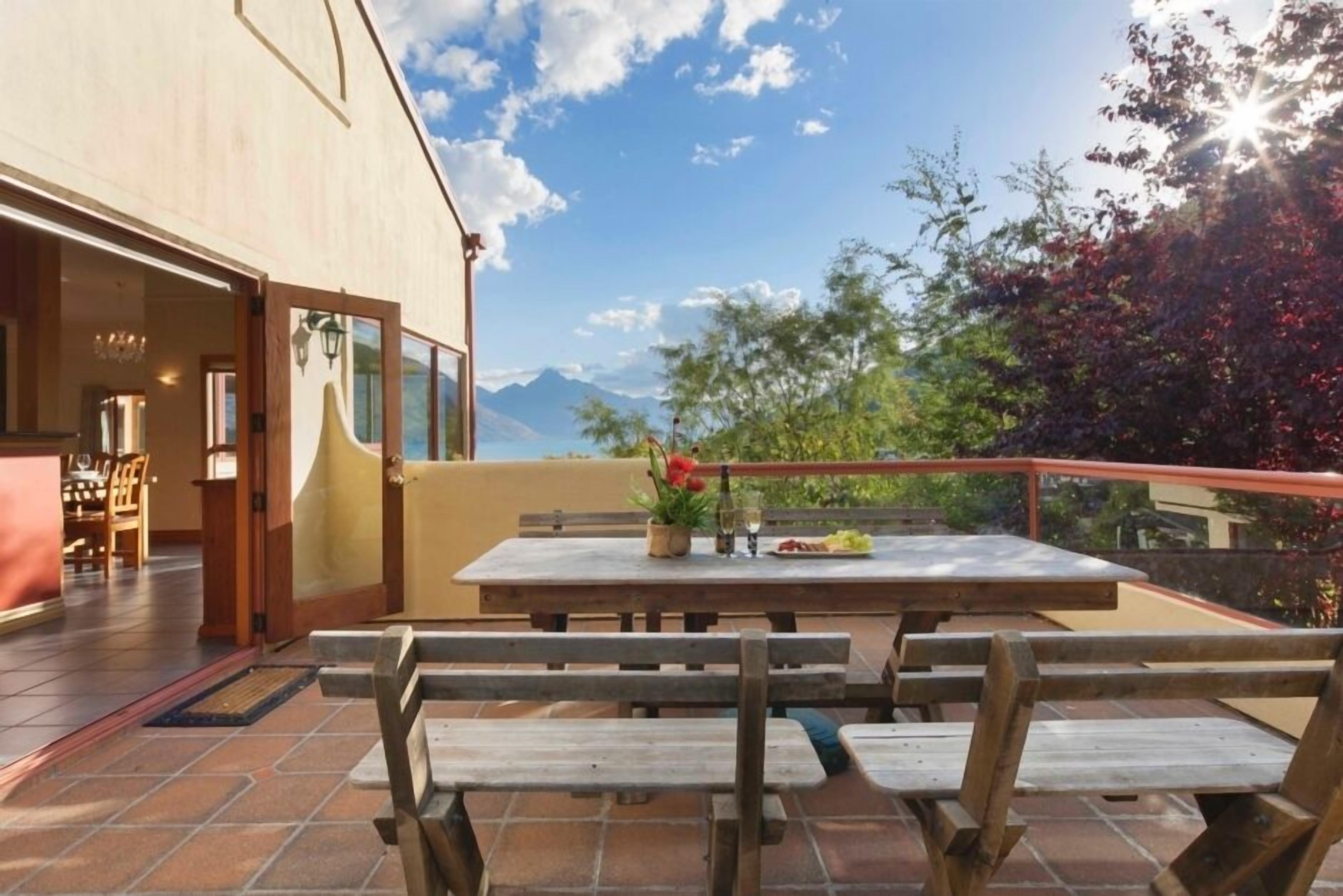 An outdoor patio with wooden benches and a table set for dining, overlooking a scenic landscape with mountains and trees.
