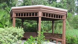 A wooden gazebo with a bench inside, surrounded by lush greenery and plants.