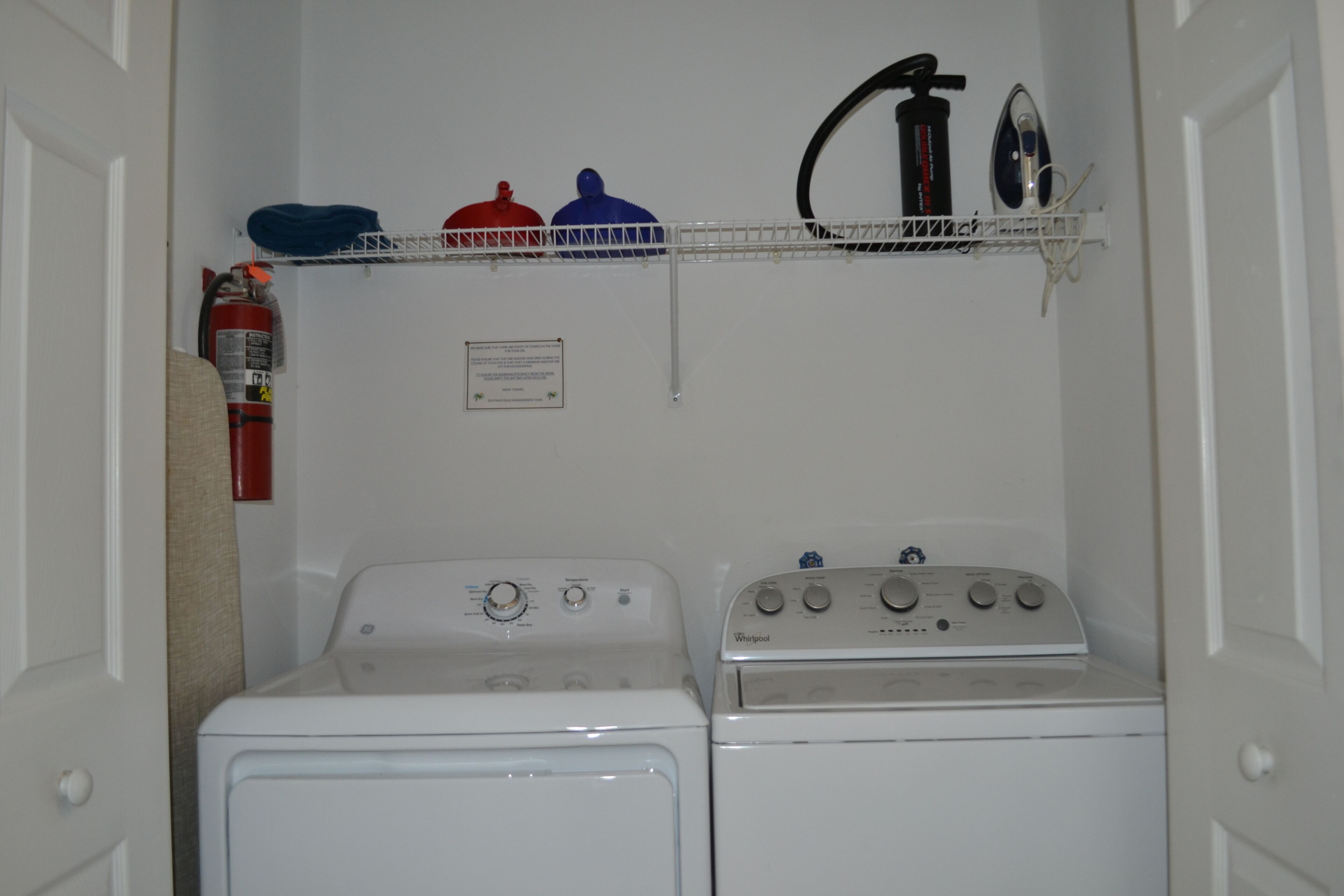 A laundry room with two washers and dryers, a fire extinguisher, and a shelf with towels and a fire hose.
