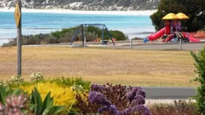Ein Spielplatz mit Rutsche und Schaukeln, ein gelber Sonnenschirm und im Hintergrund Meer und felsige Hügel.