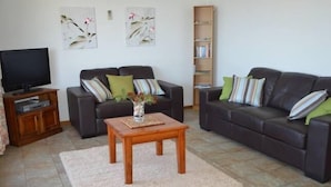 Ein Wohnzimmer mit zwei dunklen Ledersofas, einem Holztisch, einem Fernseher auf einem Holzständer und einem Bücherregal.