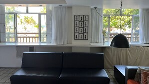 A modern living room with a black leather sofa, a black armchair, and a dining table. There are large windows with white curtains and a view of palm trees and a beach.