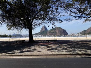 A beach with a large tree, benches, and a view of mountains in the background. - In front of the sea! Enjoy Rio with Tranquility, Comfort and many Facilities!!! (Rio de Janeiro)