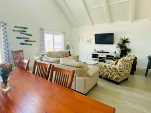 A spacious living room with a wooden dining table, beige sofas, and a patterned armchair. A flat-screen TV is mounted on the wall, and there is a potted plant in the corner. - Moondancer - Beach views, spacious , pet friendly! (Surfside Beach)