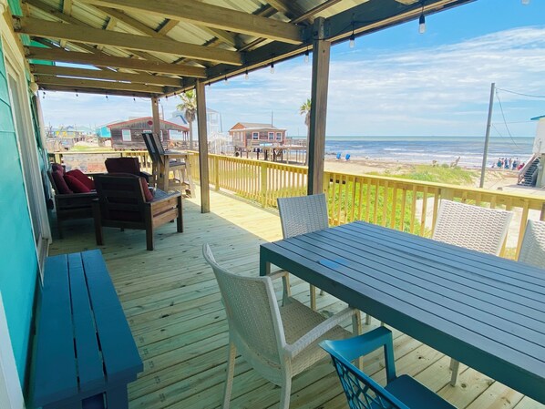 A wooden deck with a table and chairs, overlooking a beach with buildings and a clear sky. - Location, Location, Location! (Surfside Beach)