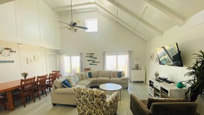 A spacious living room with a high ceiling, a ceiling fan, a large window, a sofa, a coffee table, and a TV mounted on the wall. - Location, Location, Location! (Surfside Beach)