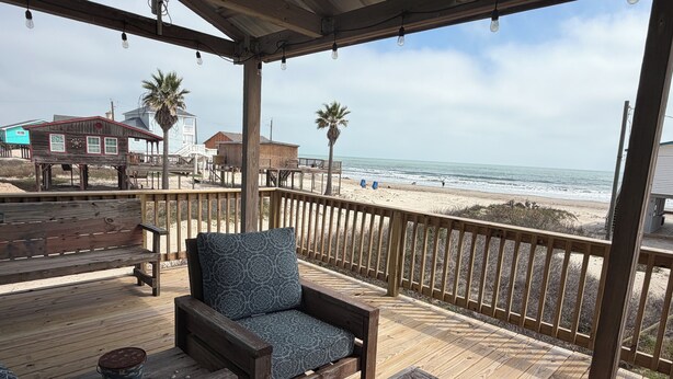 A wooden deck with a bench and a table, overlooking a beach with palm trees and a wooden structure in the background. - Location, Location, Location! (Surfside Beach)