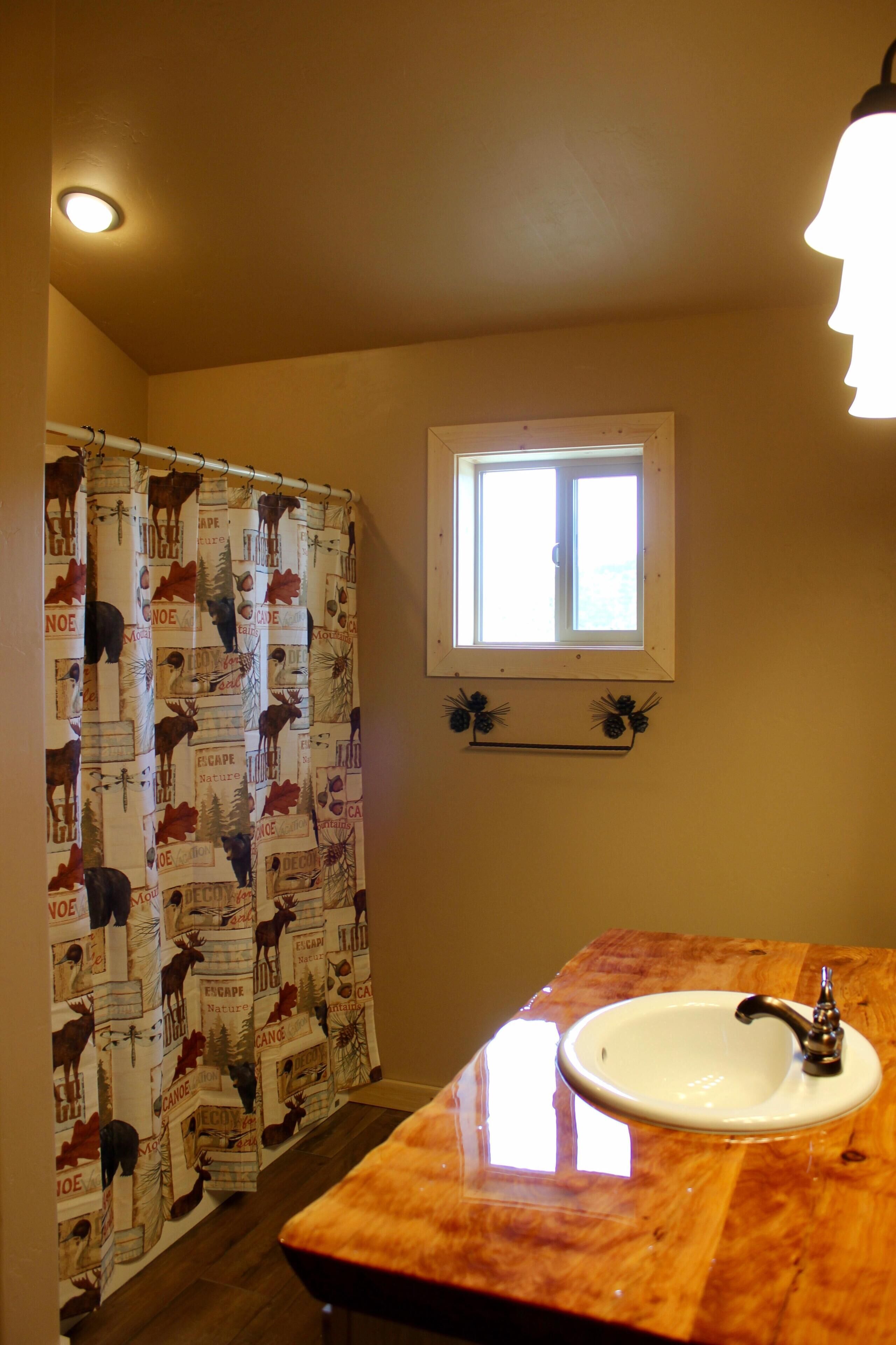 Baño con cortina de ducha estampada con motivos naturales, una ventana y un lavabo con encimera de madera.