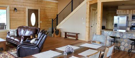 A spacious living room with a wooden floor, leather sofa, and a dining area with a large table and chairs. A staircase leads to an upper level.