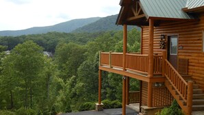 A wooden cabin with a balcony and a staircase, surrounded by lush green forest and hills.