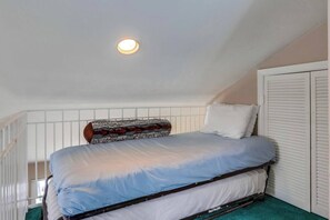 Una cama individual con cabecera, paredes blancas, una lámpara en el techo y una ventana con persianas.