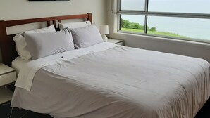 Een net opgemaakt bed met witte lakens, een houten hoofdbord en een nachtkastje met een lamp. Een raam toont uitzicht op de zee en begroeiing.