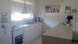Een kleine keuken met witte kastjes, een fornuis en een spoelbak. Er is een raam met lichte gordijnen en een schilderij aan de muur.