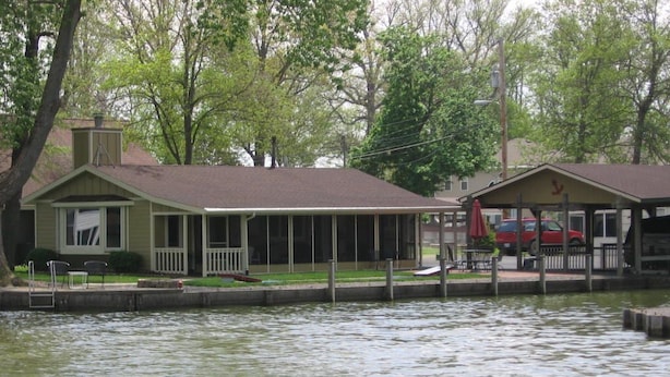 Un chalet au bord du lac avec un quai, un espace couvert où se trouve une voiture rouge, et une terrasse enherbée.
