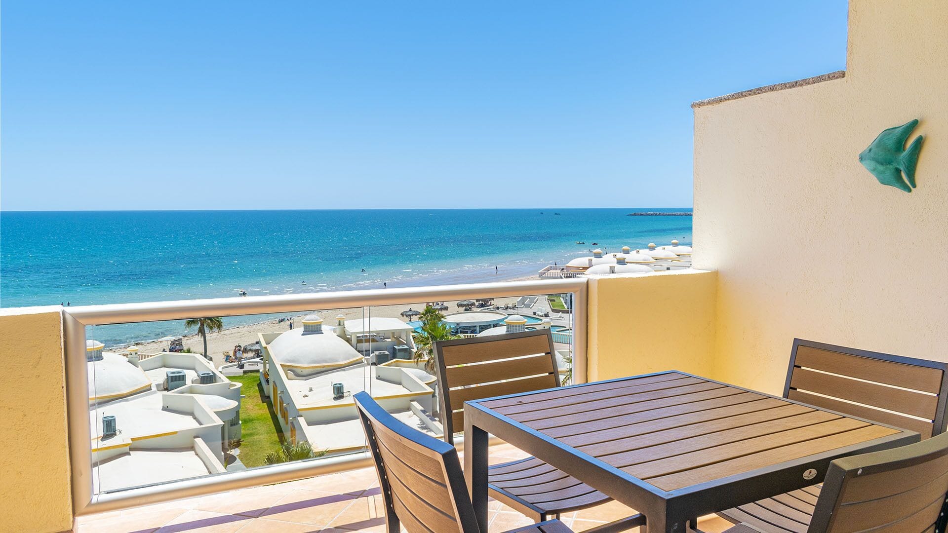 Un balcon avec une table et des chaises en bois, donnant sur une plage parsemée de parasols et une piscine.