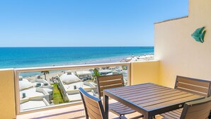 Un balcón con mesa y sillas de madera, con vista a una playa con sombrillas y una piscina.