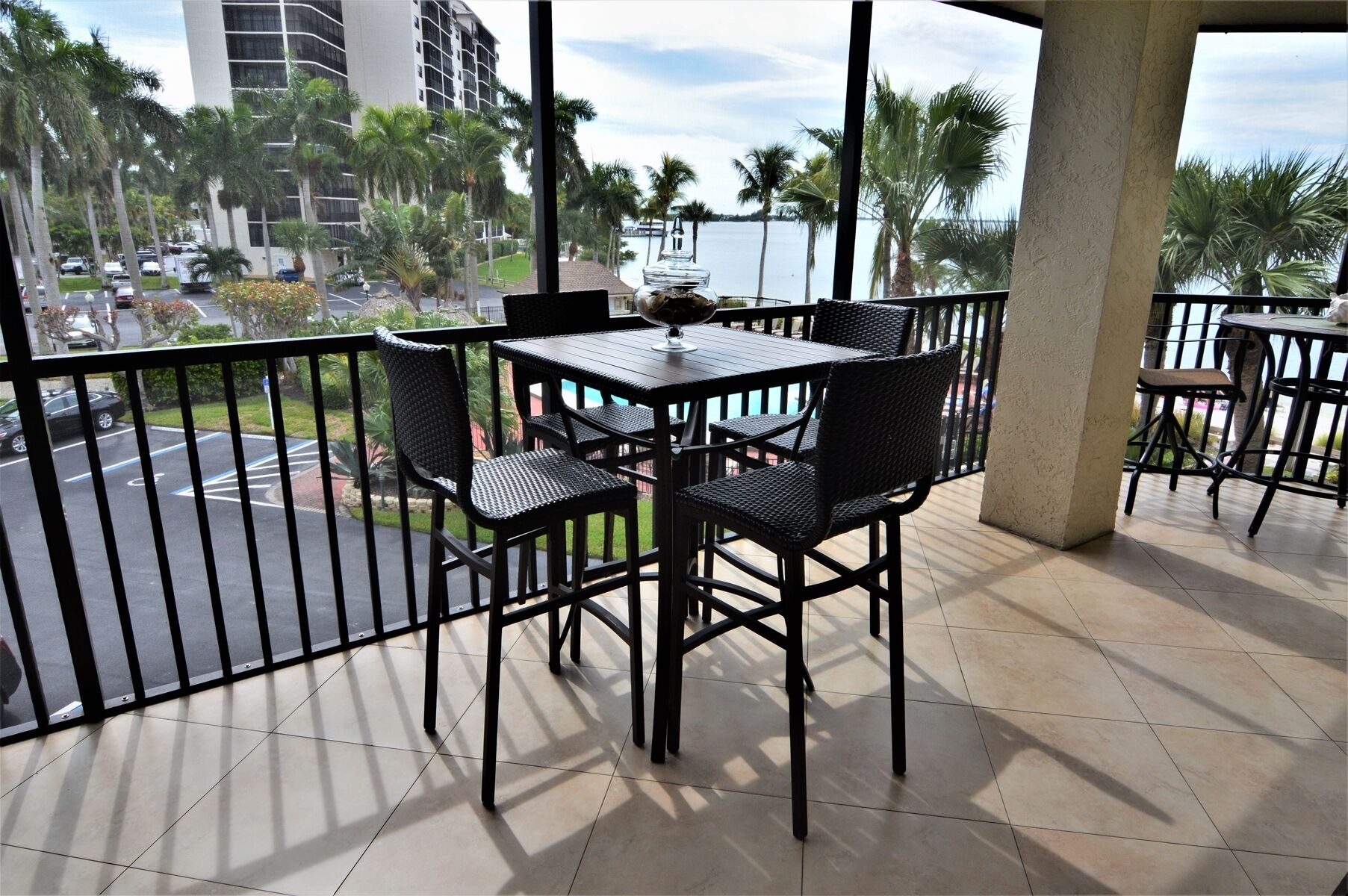 A balcony with a table and chairs, overlooking a parking area and a body of water with palm trees.