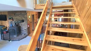 Eine Holztreppe mit Geländer führt zu einem Bereich im zweiten Stock. Im ersten Stock ist eine kleine Holzplattform mit einer Vogel-Skulptur zu sehen.