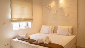 A bedroom with a large bed, a smaller bed, a window with blinds, and a wooden headboard with wall-mounted lights.